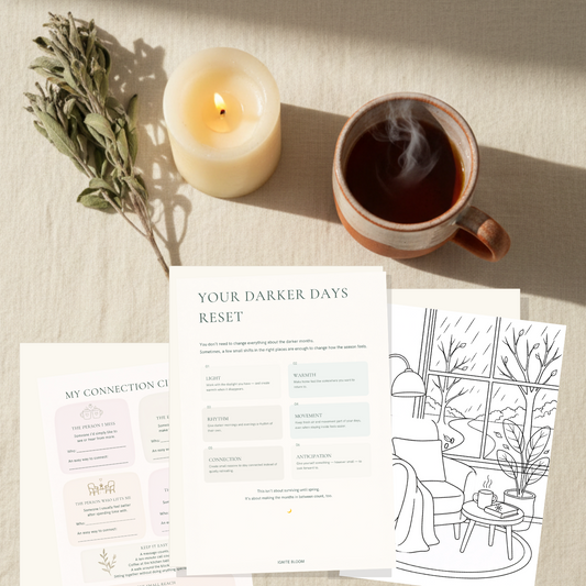 A styled flat-lay of printed journaling or therapy worksheets titled 'YOUR DARKER DAYS RESET' and other matching pages, arranged on a light fabric surface beside a lit candle, dried herbs, and a cup of tea.