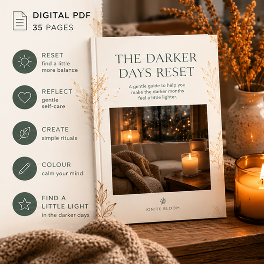 Digital product mockup for a PDF guide titled 'THE DARKER DAYS RESET' by Ignite Bloom, shown as a book cover standing on a wooden surface with candles and dried flowers. A vertical sidebar on the left lists five benefits in green icons and text: reset, reflect, create simple rituals, colour, and find a little light.